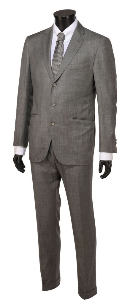 A full-length view of a gray men's suit displayed on a mannequin. The suit includes a blazer with a notched lapel, dressed shirt, and a patterned tie.